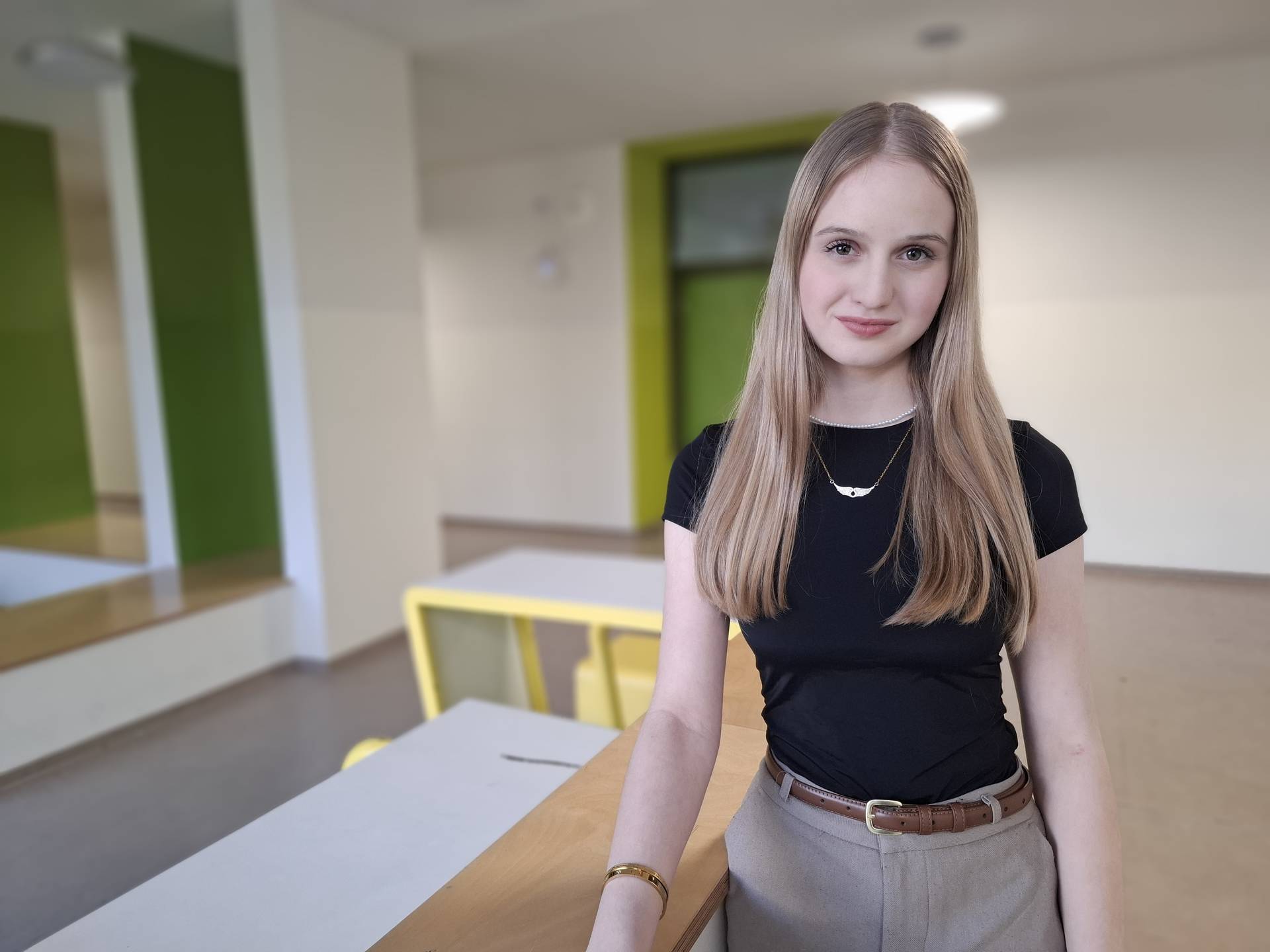 Junge Frau mit langen Haaren, schwarzem Top und beige Hose, lächelt in einem hellen Raum mit grünen Akzenten